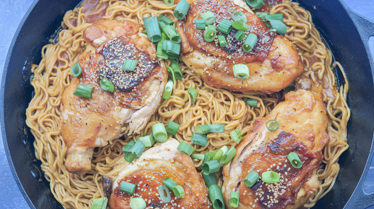 A rustic skillet filled with golden-seared chicken breasts placed on a bed of buttery, Parmesan-infused egg noodles, garnished with fresh parsley, sitting on a wooden table.