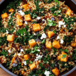 A rustic black cast-iron pan filled with a vibrant quinoa and kale pilaf, garnished with roasted butternut squash, crumbled feta cheese, and fresh herbs.