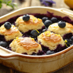 Golden biscuits swimming in purple blueberry sauce in a cream ceramic dish, garnished with fresh blueberries and lemon zest on a rustic wooden table.