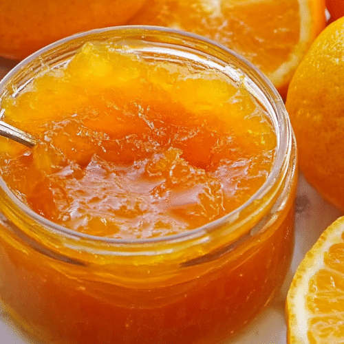 Homemade orange and lemon marmalade in a glass jar with a silver spoon, surrounded by fresh citrus fruits on a marble surface.